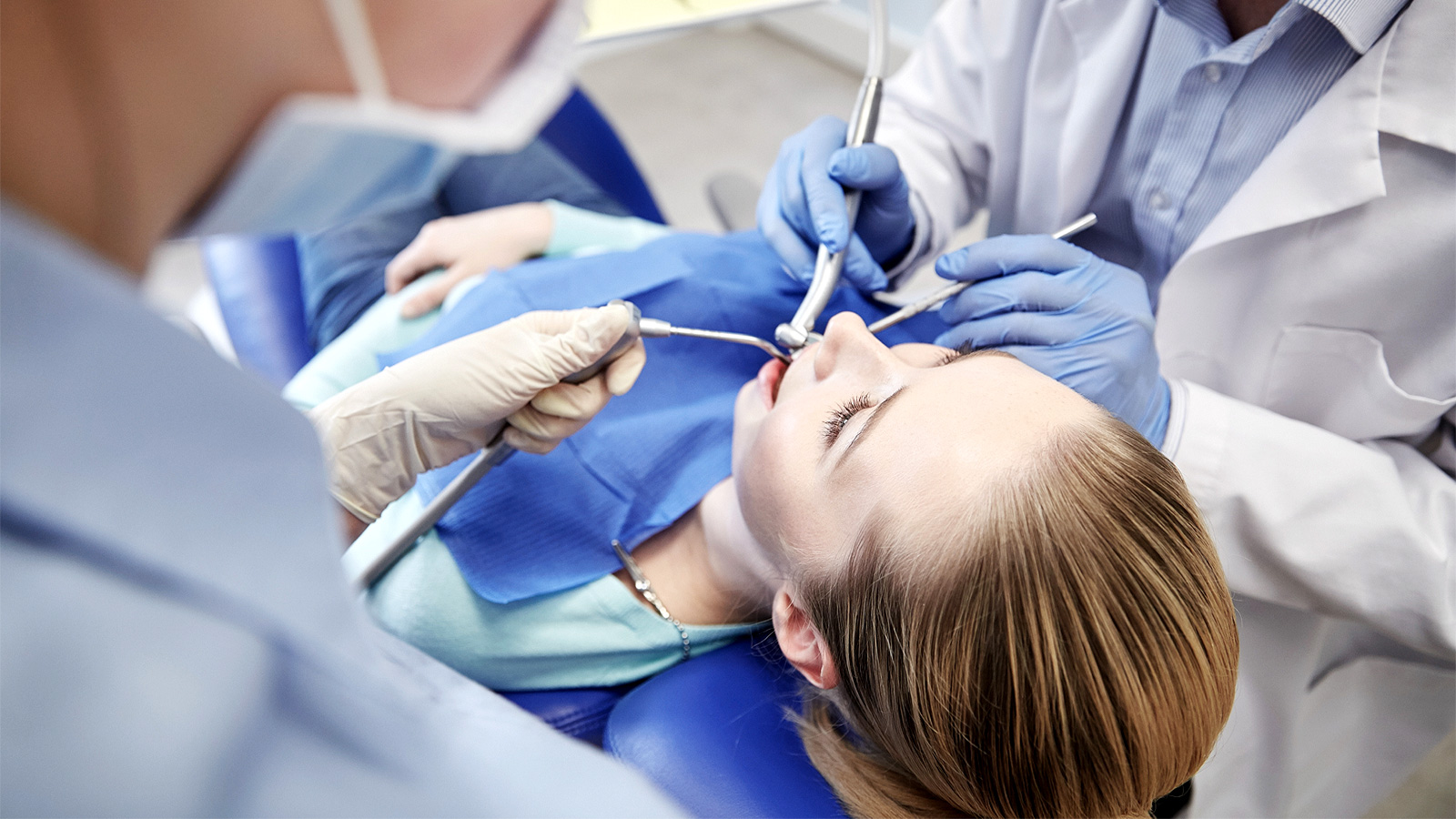 The dentist and assistant treating the patient’s teeth using a mirror, handpiece and dental air syringe in a dental clinic.